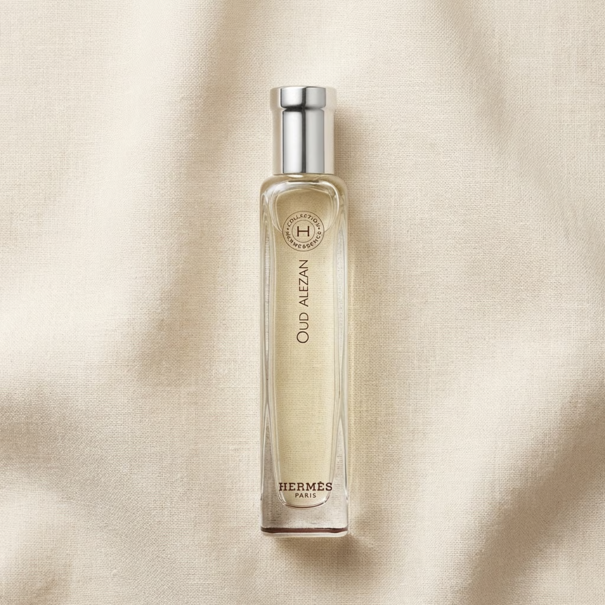 A bottle of Hermes Oud Alzéan perfume, featuring a clear glass rectangular bottle with a silver cap, filled with a pale golden liquid, set against a draped beige fabric background.