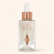 Charlotte's Magic Serum Crystal Elixir in a clear glass dropper bottle with a rose gold collar and white dropper cap, set against a solid pale beige background.