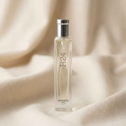A clear rectangular perfume bottle with a silver cap, standing upright on draped cream fabric. The bottle is transparent and shows the liquid inside, with 'F24' and 'HERMES PARIS' printed on the front.