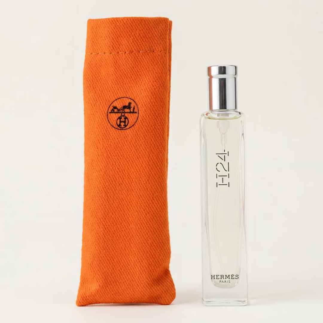 A clear rectangular Hermes H24 perfume bottle with a silver cap stands next to an orange canvas pouch featuring the black Hermes logo, on a light beige background.